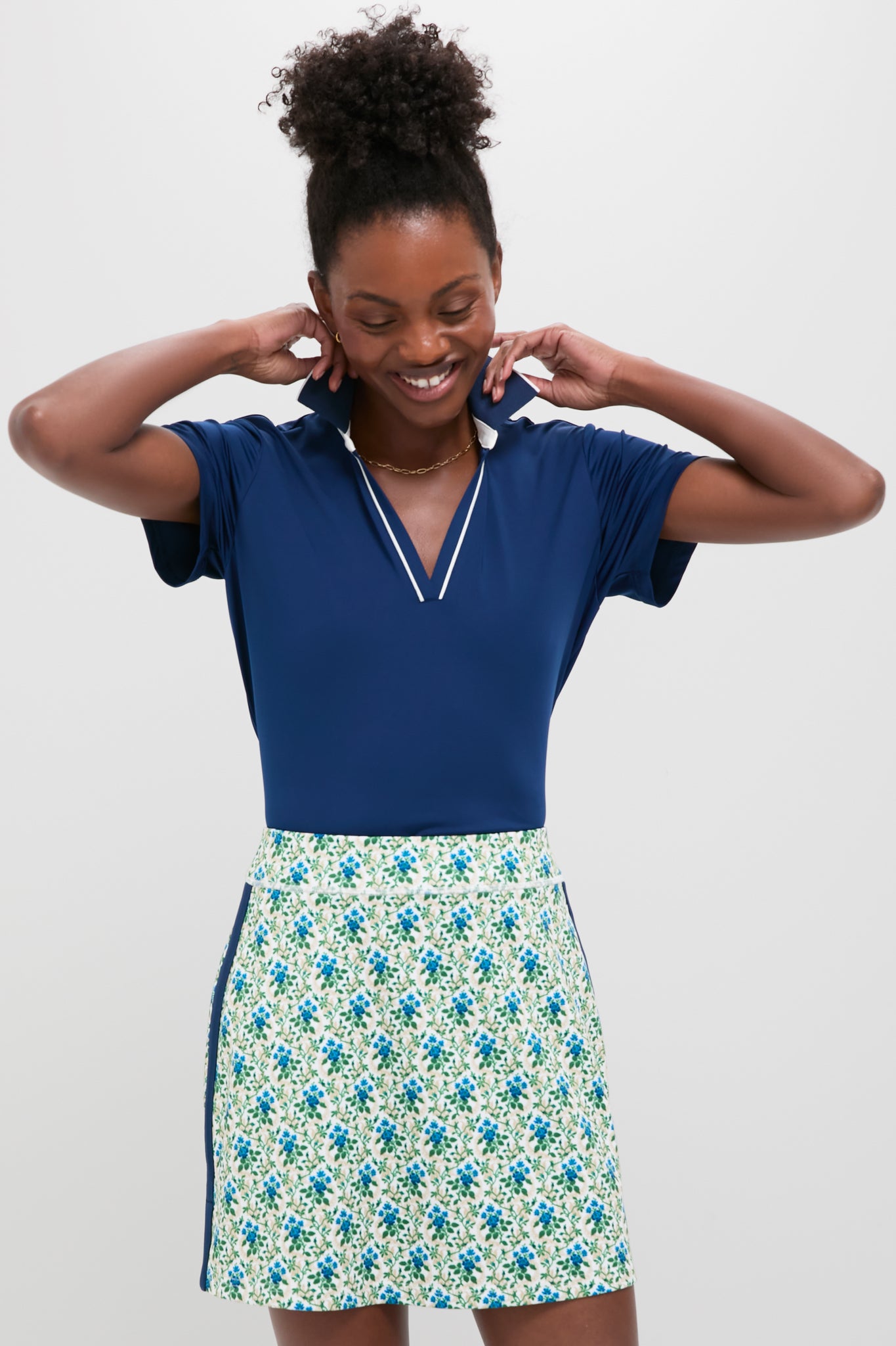 A woman smiles, wearing a Tnuck Sport navy Short Sleeve Polo with SPF Protection and a white skirt featuring a blue and green floral print.