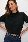 A woman stands against a white background wearing a Tuckernuck black button-front cardigan, blue jeans, a snake-print belt, and gold earrings.