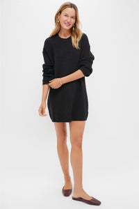 A woman smiles against a white background wearing a Pomander Place long-sleeve black sweater dress with a mod silhouette and brown flats, her arm bent at the elbow.