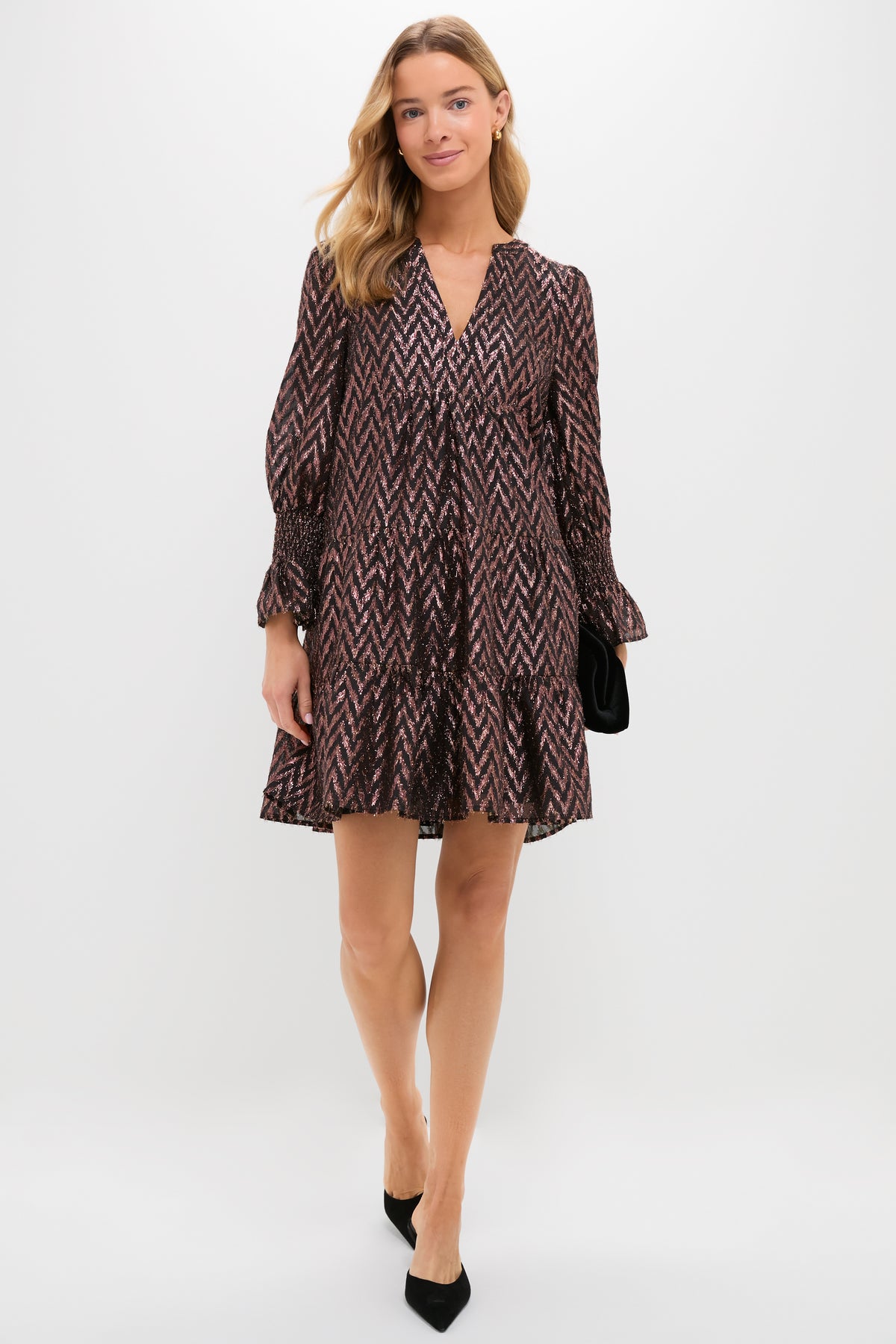 A woman in a loose, patterned Metallic Ikat V-neck dress by Pomander Place, paired with black heels and a black clutch, stands against a plain white background.