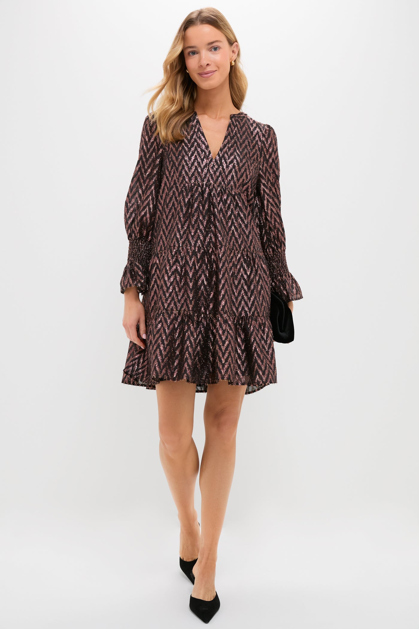 A woman in a loose, patterned Metallic Ikat V-neck dress by Pomander Place, paired with black heels and a black clutch, stands against a plain white background.