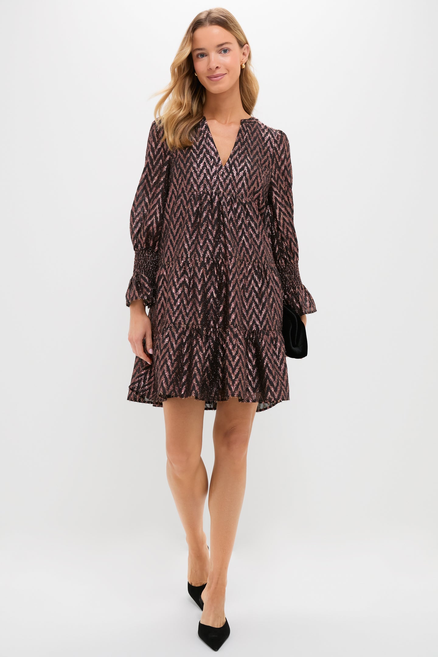 A woman in a loose, patterned Metallic Ikat V-neck dress by Pomander Place, paired with black heels and a black clutch, stands against a plain white background.