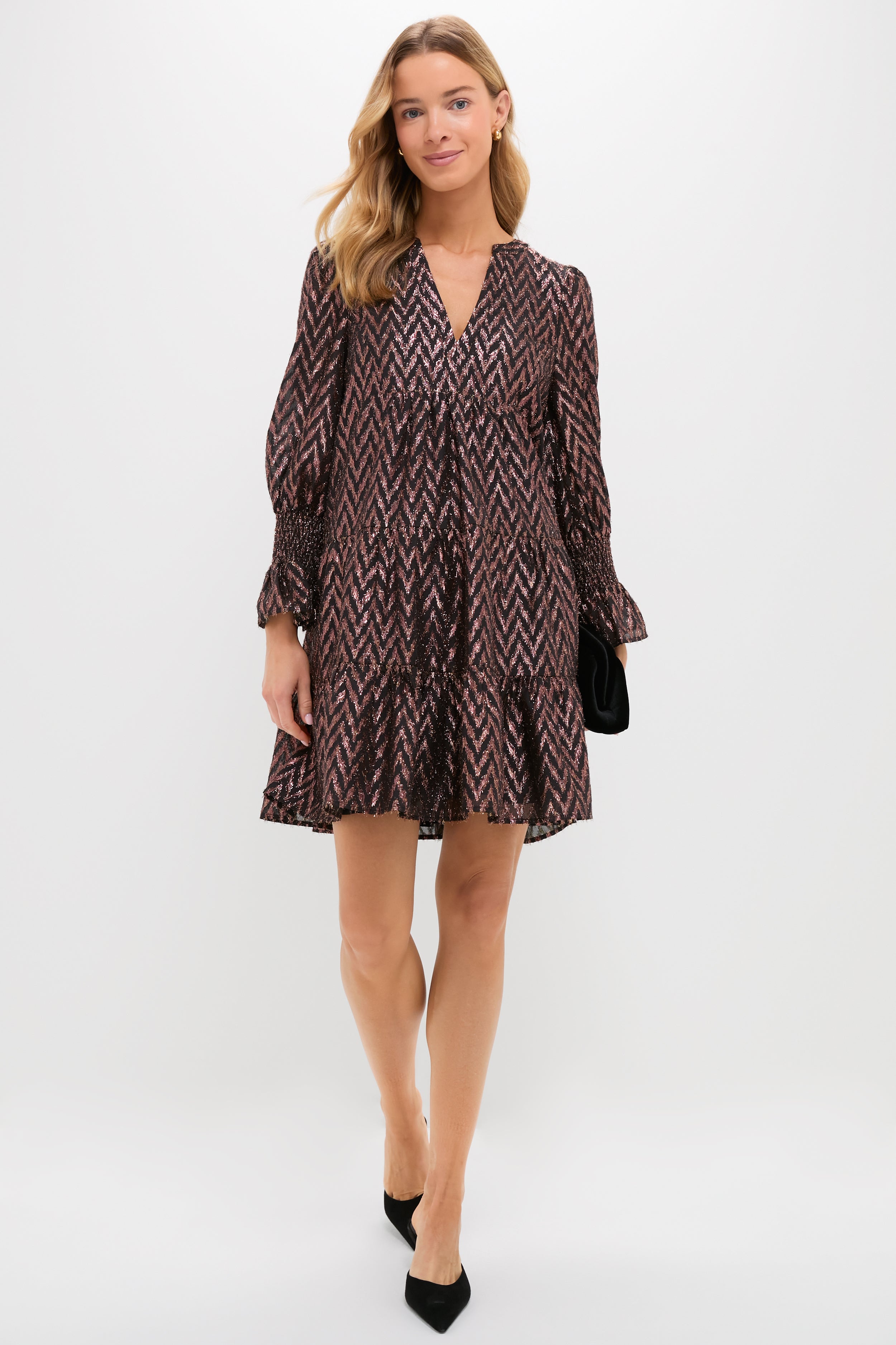 A woman in a loose, patterned Metallic Ikat V-neck dress by Pomander Place, paired with black heels and a black clutch, stands against a plain white background.