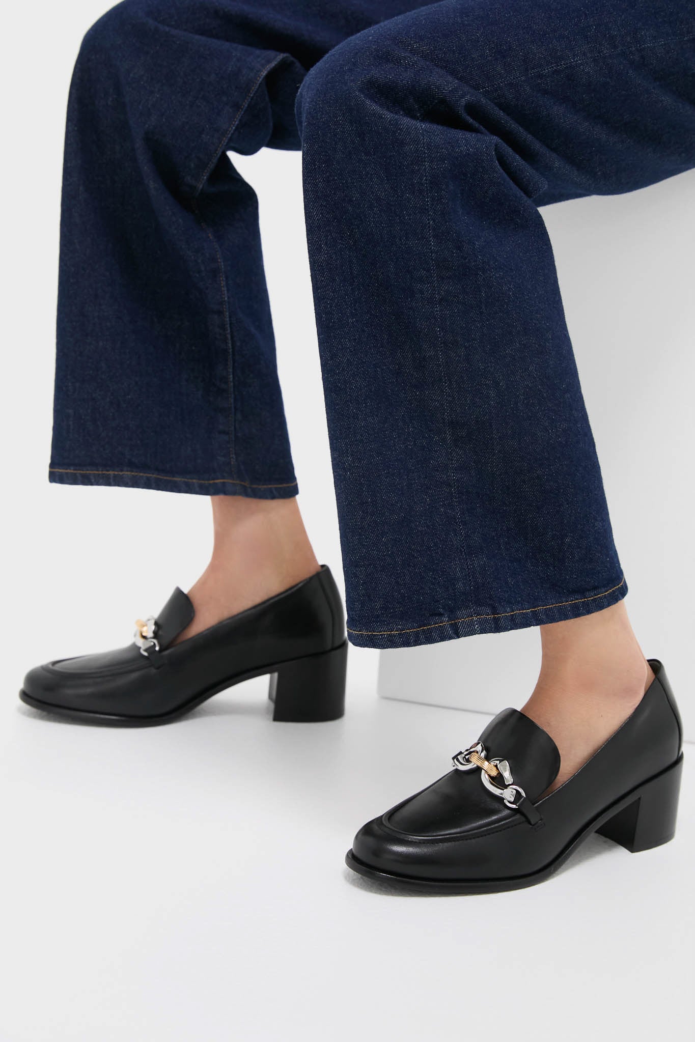 A person sits on a white surface wearing dark blue cropped jeans and Tory Burch black heeled loafers featuring a buckle detail.