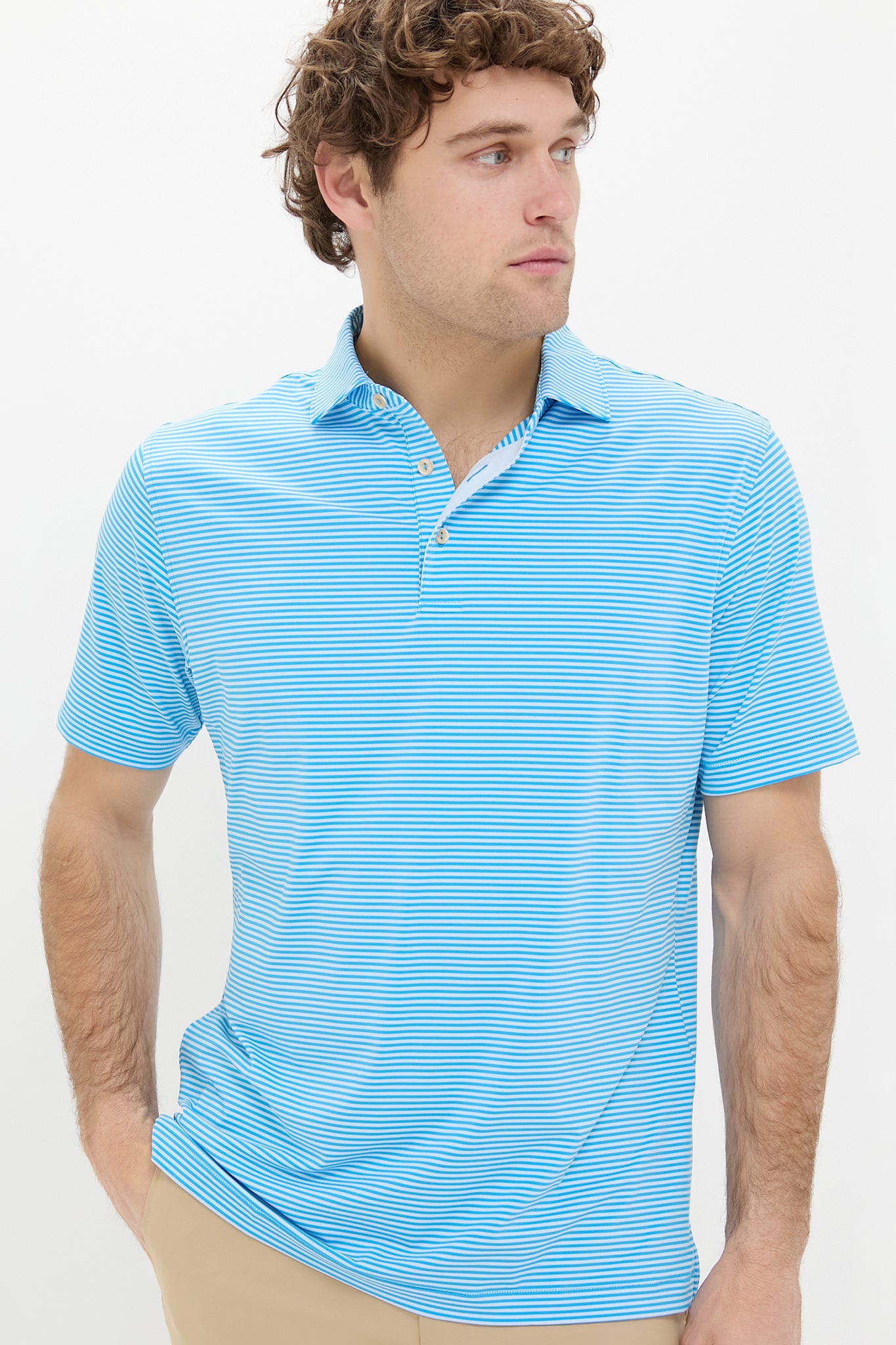 A man confidently wears a Peter Millar light blue and white striped performance jersey polo with four-way stretch, paired with tan pants.