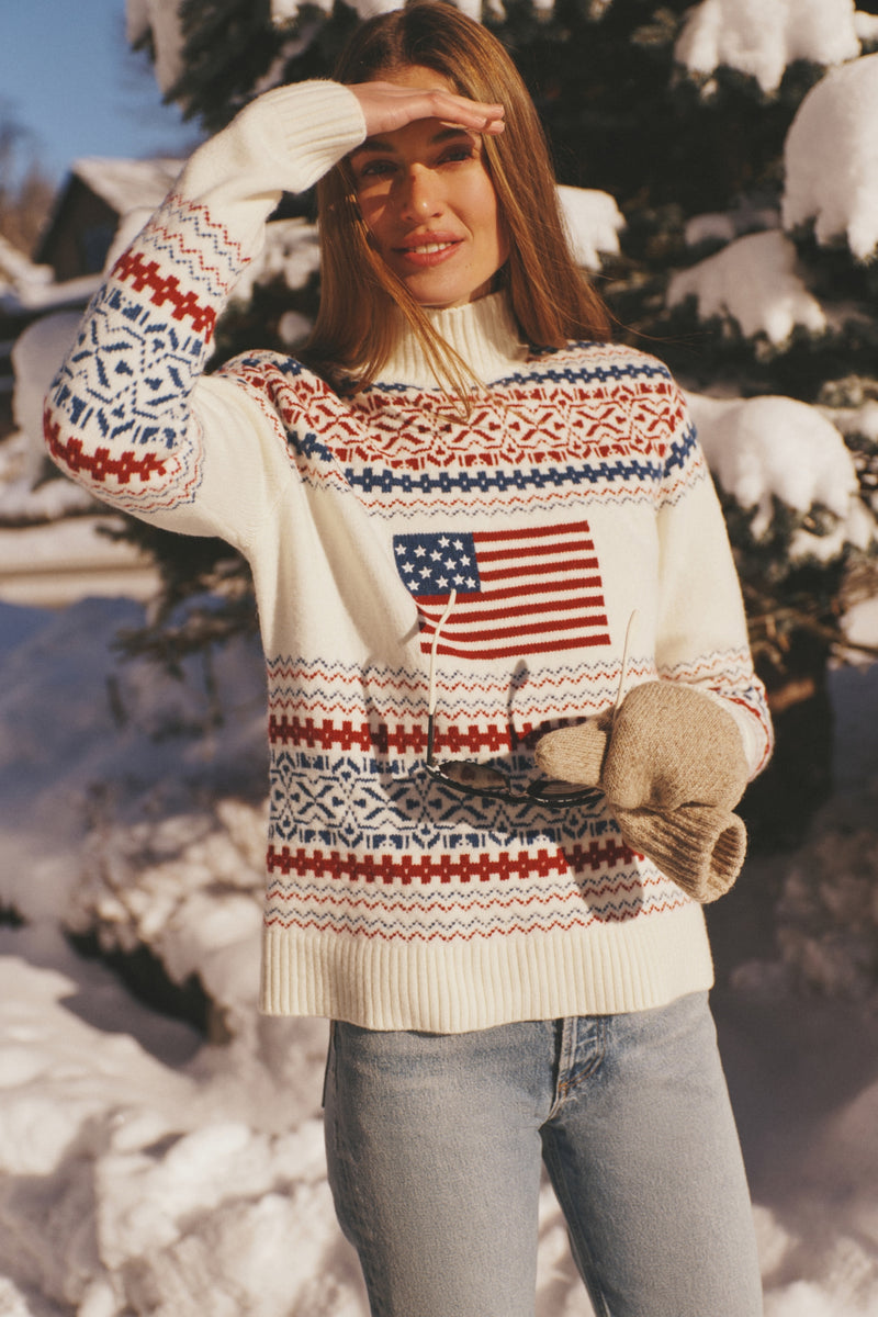 Young woman in snowy landscape wearing the classic, high-end American Flag Christie Turtleneck Sweater, effortlessly styled with light jeans and holding beige mittens.