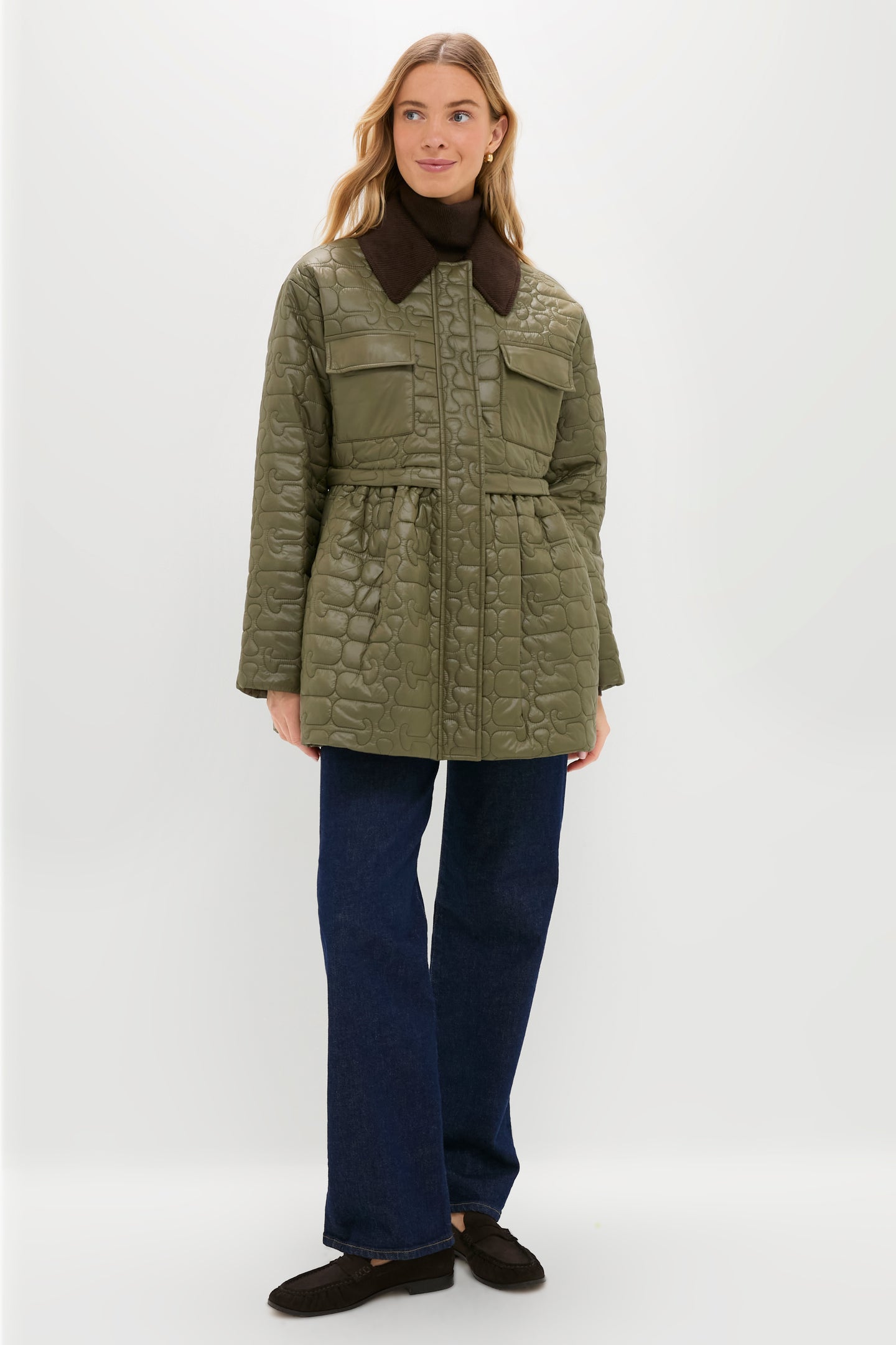 A woman stands against a plain white background wearing a green GANNI quilted jacket with front pockets and a dark collar, paired with blue jeans and black shoes.