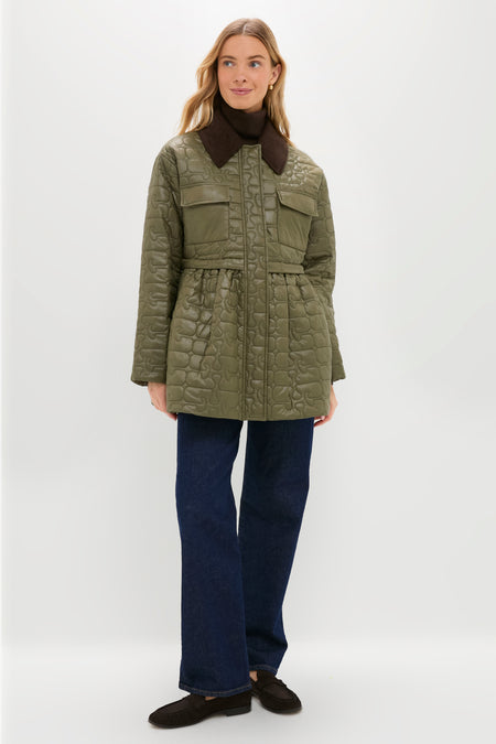 A woman stands against a plain white background wearing a green GANNI quilted jacket with front pockets and a dark collar, paired with blue jeans and black shoes.