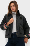 Woman in a black Barbour x Tuckernuck Kinsley diamond quilted jacket with floral lining, light gray turtleneck, and dark blue jeans stands against a plain white background.