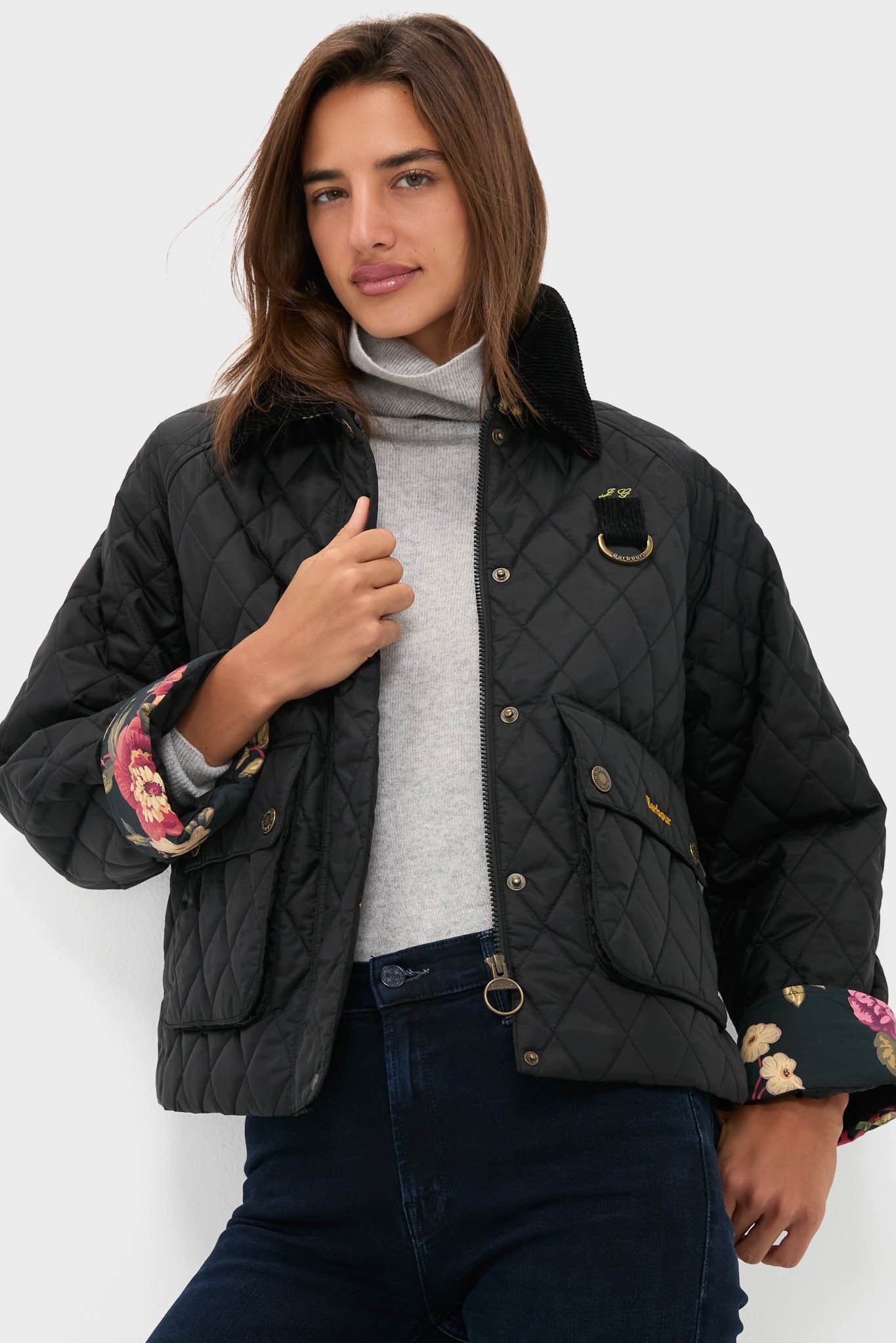 Woman in a black Barbour x Tuckernuck Kinsley diamond quilted jacket with floral lining, light gray turtleneck, and dark blue jeans stands against a plain white background.