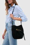 Person with classic Black Suede Belted Bucket Bag, featuring gold-tone buckle, effortlessly styled with light-blue shirt and jeans, embodying fun, high-end fashion.