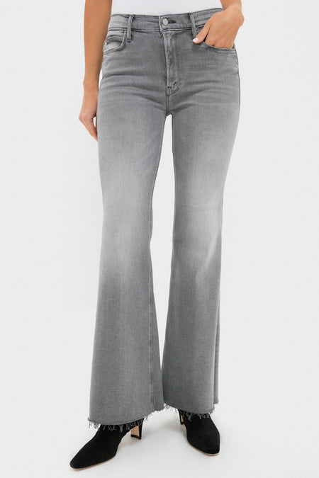 A person wears high-waisted, gray MOTHER jeans with a flared cut and frayed hems, paired with black ankle boots, standing against a plain white background.