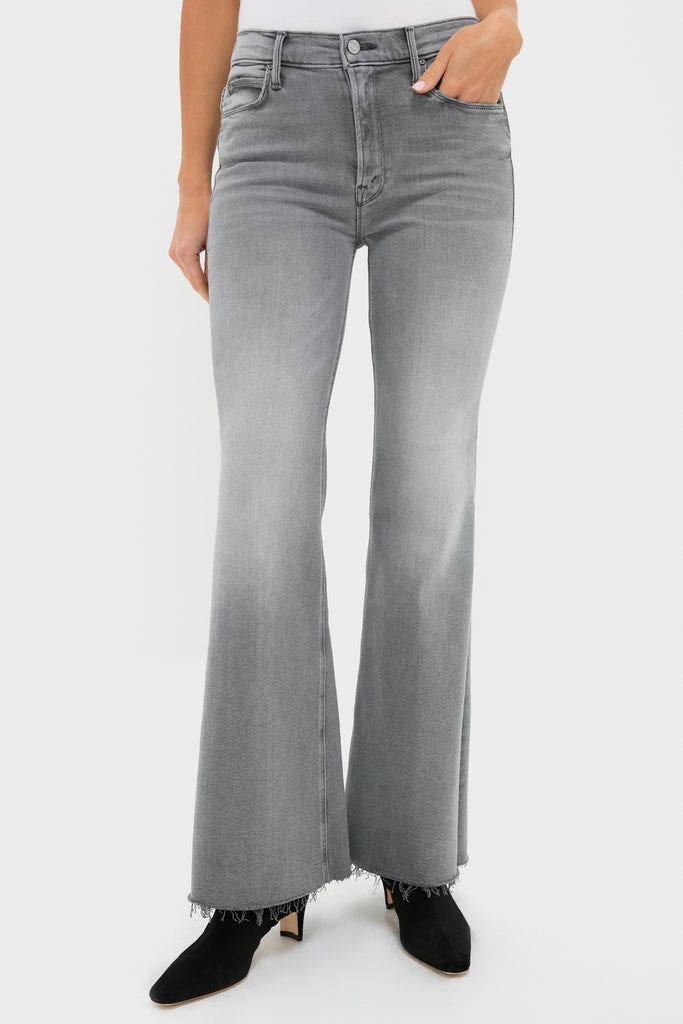 A person wears high-waisted, gray MOTHER jeans with a flared cut and frayed hems, paired with black ankle boots, standing against a plain white background.