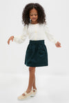 Young girl models the classic Navy Douchka Skirt, featuring heart-shaped pockets and an elastic waist, paired with a cream cardigan and glittery Mary Jane flats for an effortless, high-end look.