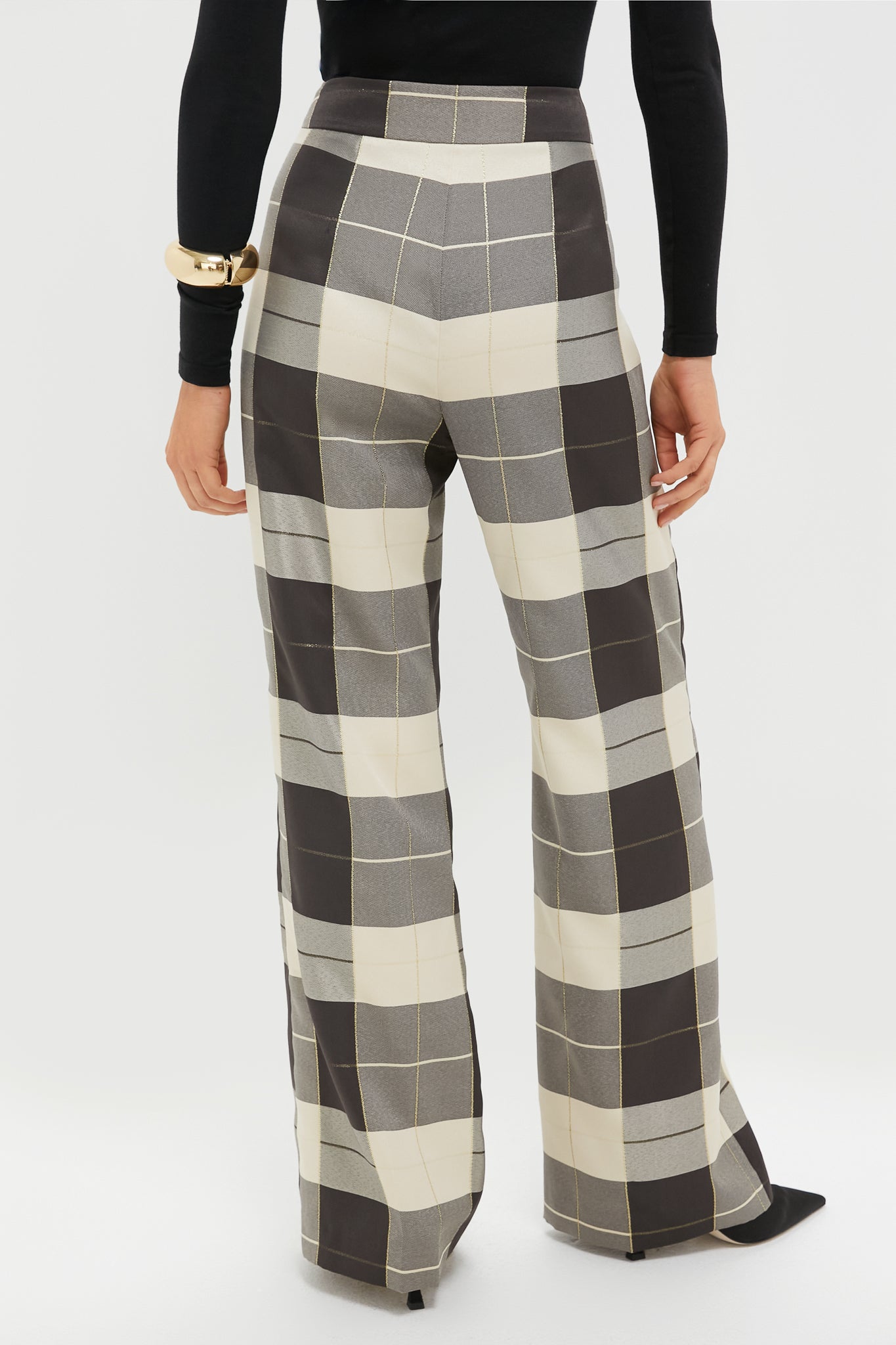 A person wearing Tuckernuck black and white plaid wide-leg high-waist trousers, paired with a black long-sleeve top and chunky gold bracelet, seen from the back.