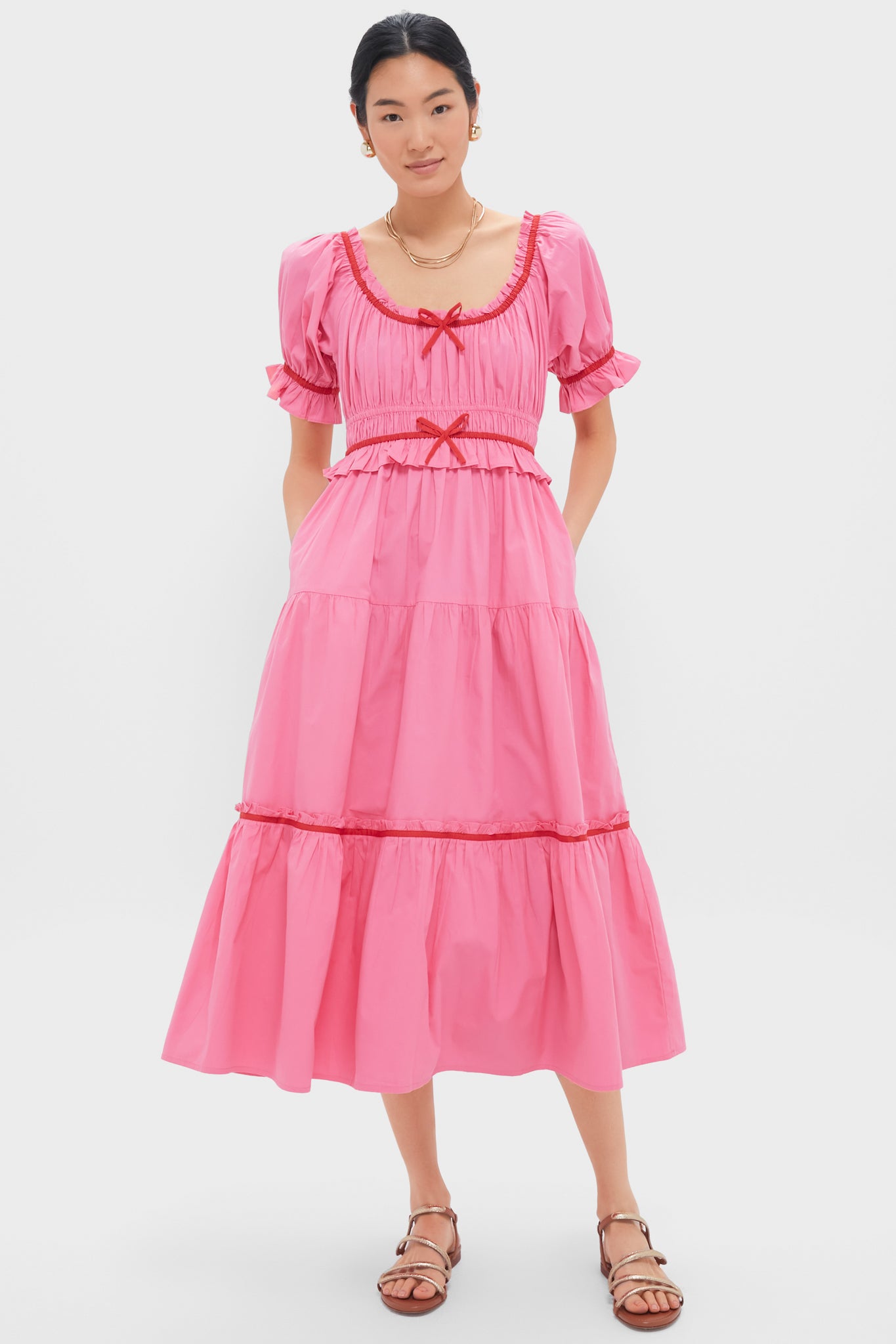 A woman wears a Pink City Prints pink organic cotton midi dress with puff sleeves, red trim, and red bows, paired with brown sandals.
