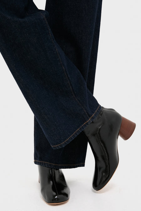 A person wears dark blue jeans and Mansur Gavriel classic leather booties, crossing one leg over the other against a plain background.