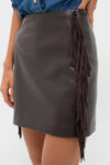 Woman wearing the Coffee Faux Leather Fringe Paddock Mini Skirt, showcasing its classic A-line silhouette and fun fringe detailing, paired effortlessly with a denim shirt.