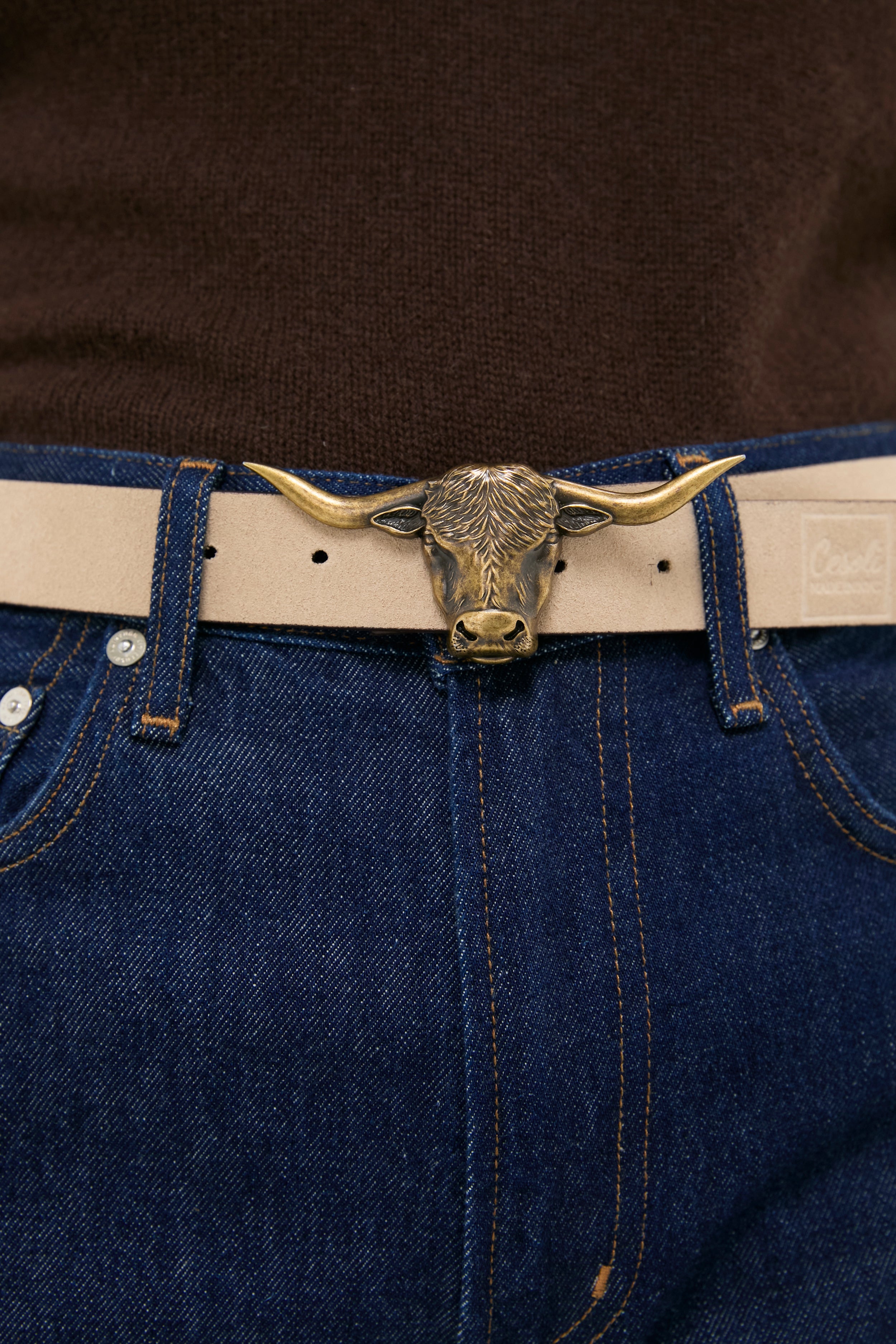 Brass Lansing Longhorn Buckle in polished brass, adorning a light-tan leather belt on dark denim jeans, offering a classic, effortless style with a high-end, fun twist.