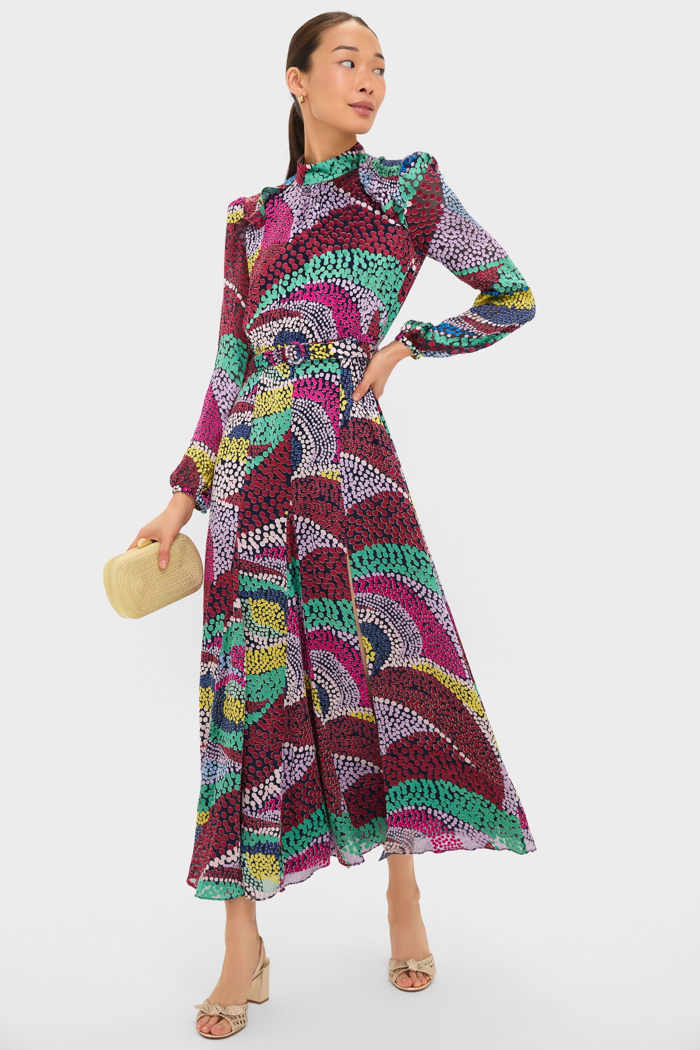 Model wearing the Jangle Jacqui-B Dress, a classic, high-end maxi with a colorful dot pattern, mock neck, and flowing skirt, styled with a woven clutch and sandals.