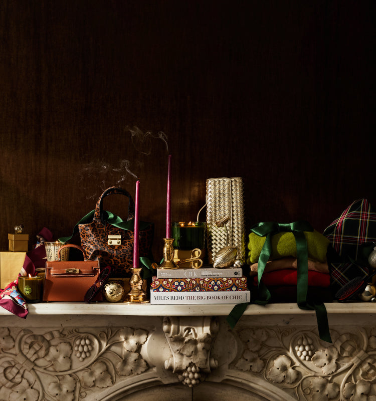 A thoughtful selection of gifts sitting on a fireplace mantel. Gifts include candles, bags, clutches, and scarves. 