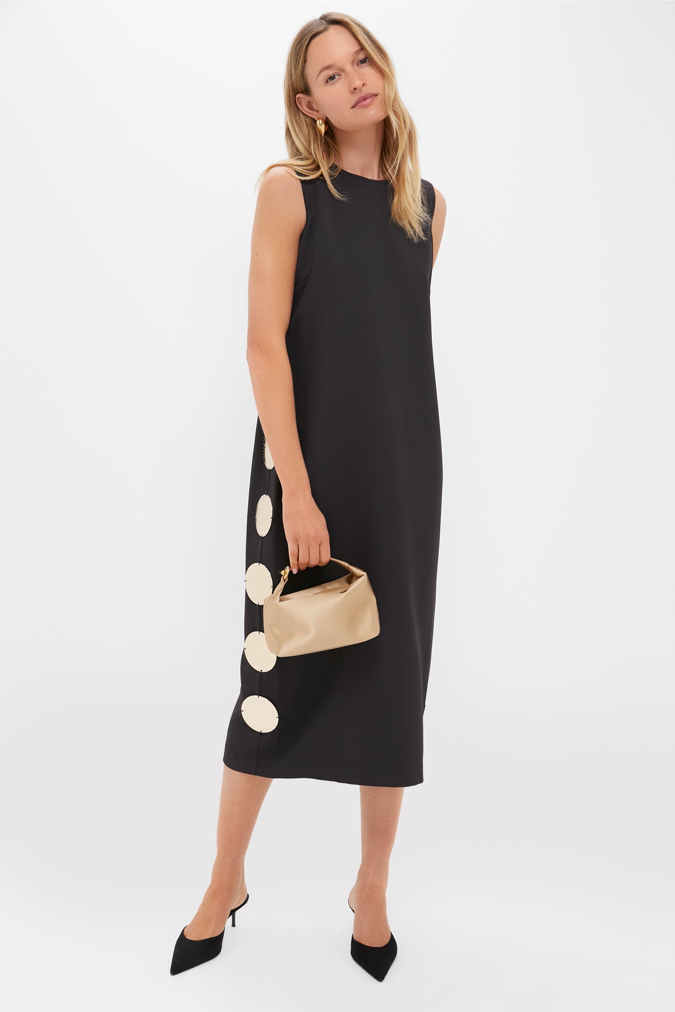 A woman in a Hyacinth House sleeveless black shift dress with large white polka dots on one side, holding a beige clutch and wearing black heels, stands against a plain background.
