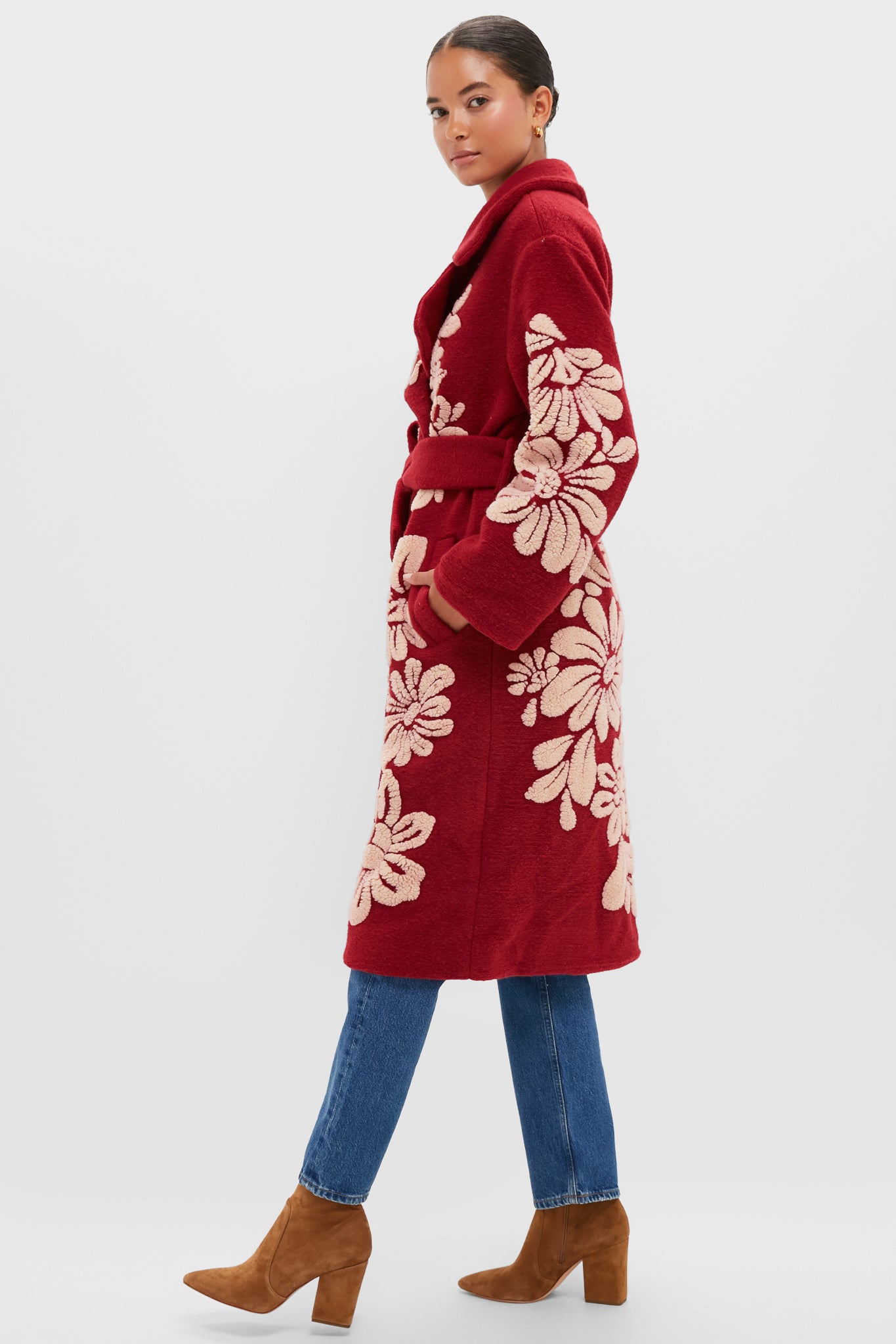 A woman wears a vibrant long red belted coat with bold floral patterns from Farm Rio, paired with blue jeans and tan suede boots, standing against a plain background.