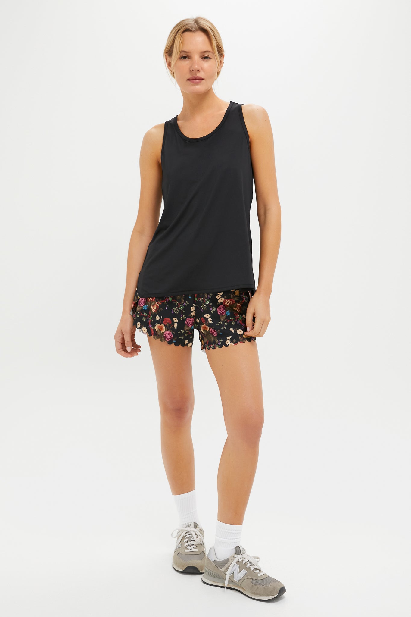 A person in a Tnuck Sport black racerback tank, black floral quick-dry shorts, white socks, and gray sneakers stands against a plain white background.