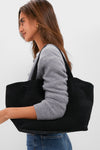 Woman carrying the Black Suede Blaine Tote, a classic, high-end bag with an effortless style, featuring top handles and a woven flap for a fun, polished look.