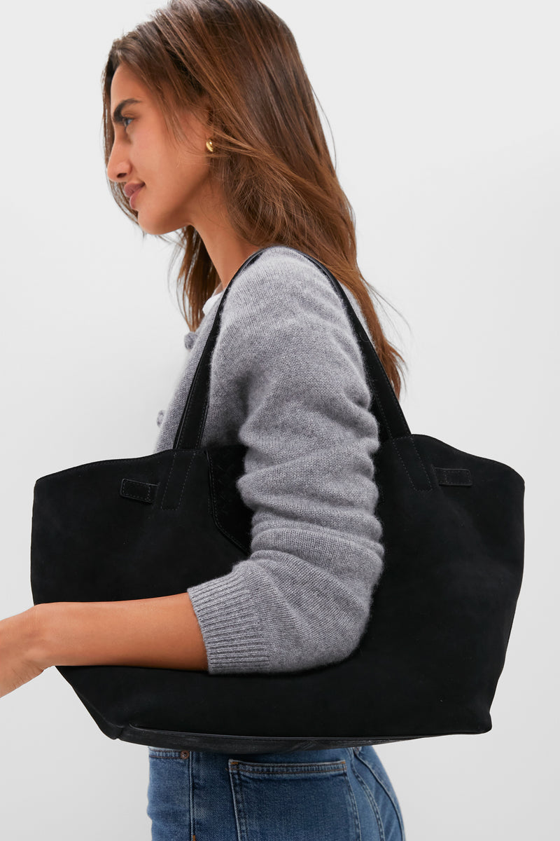 Woman carrying the Black Suede Blaine Tote, a classic, high-end bag with an effortless style, featuring top handles and a woven flap for a fun, polished look.