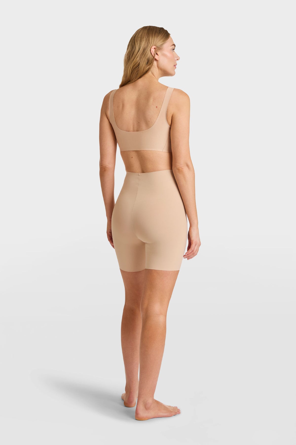 A woman stands barefoot facing away, wearing Commandos nude seamless bra and high-waisted shapewear short crafted from breathable fabric, set against a plain white background.