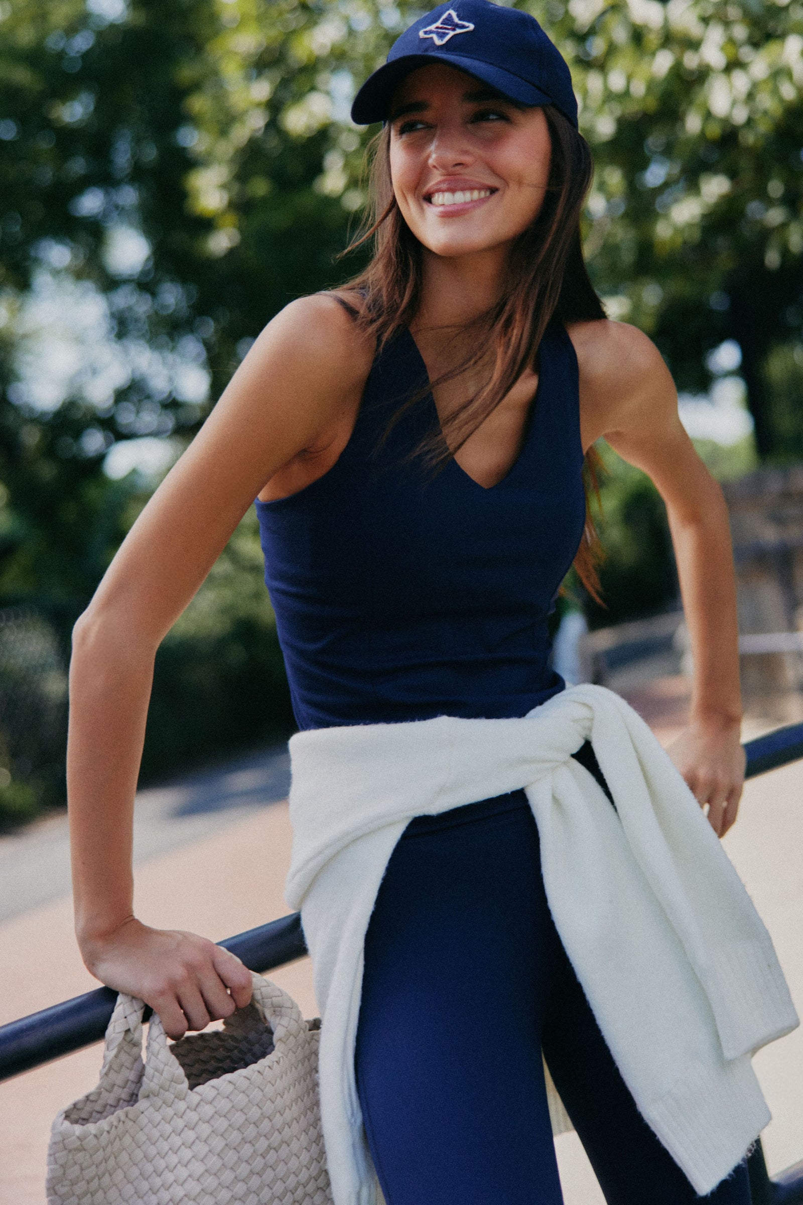 A woman wearing Tnuck Sport navy athleisure and a cap smiles outdoors, carrying a woven bag with a white sweater tied at her waist. Trees and a railing appear in the background.