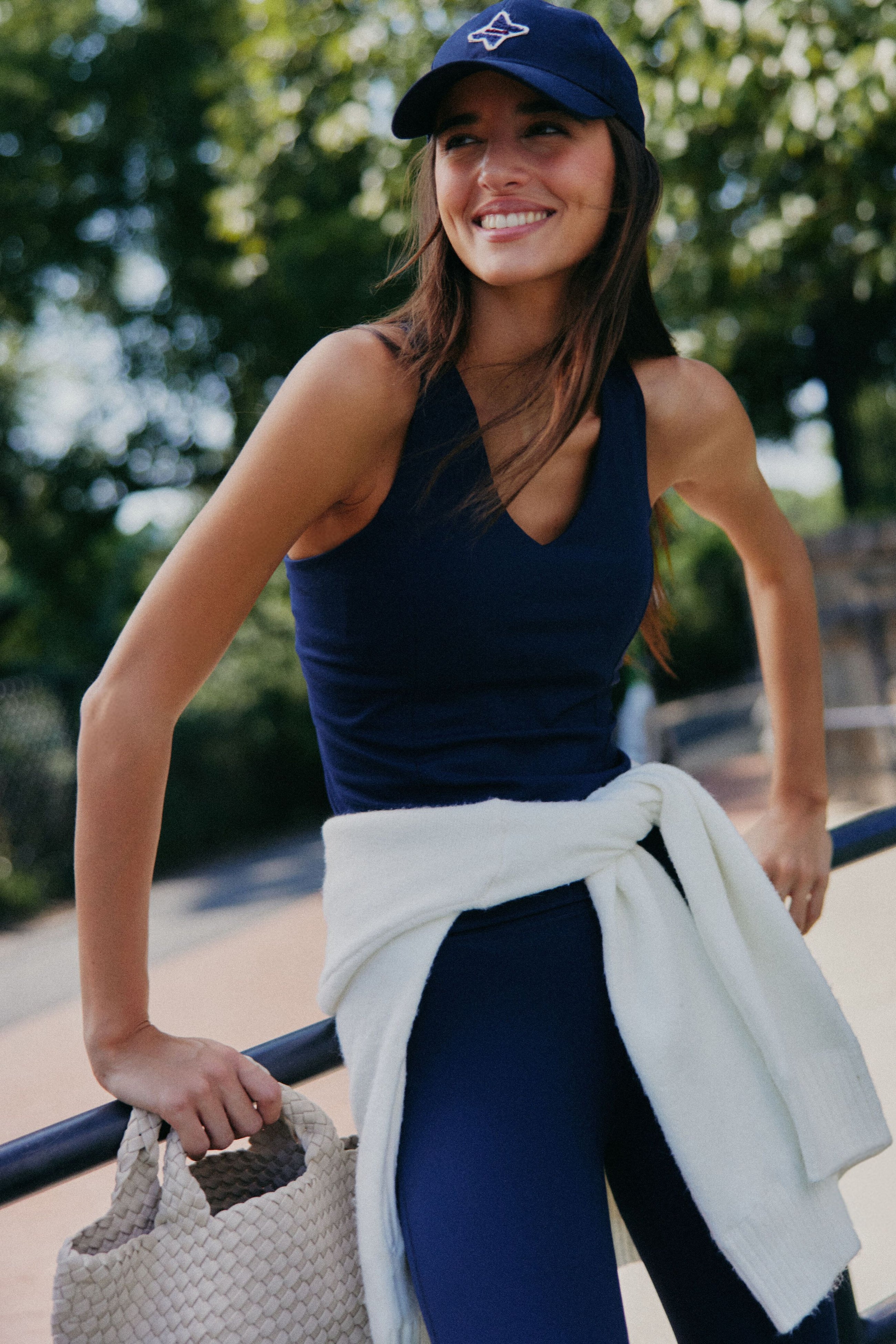 A woman wearing Tnuck Sport navy athleisure and a cap smiles outdoors, carrying a woven bag with a white sweater tied at her waist. Trees and a railing appear in the background.