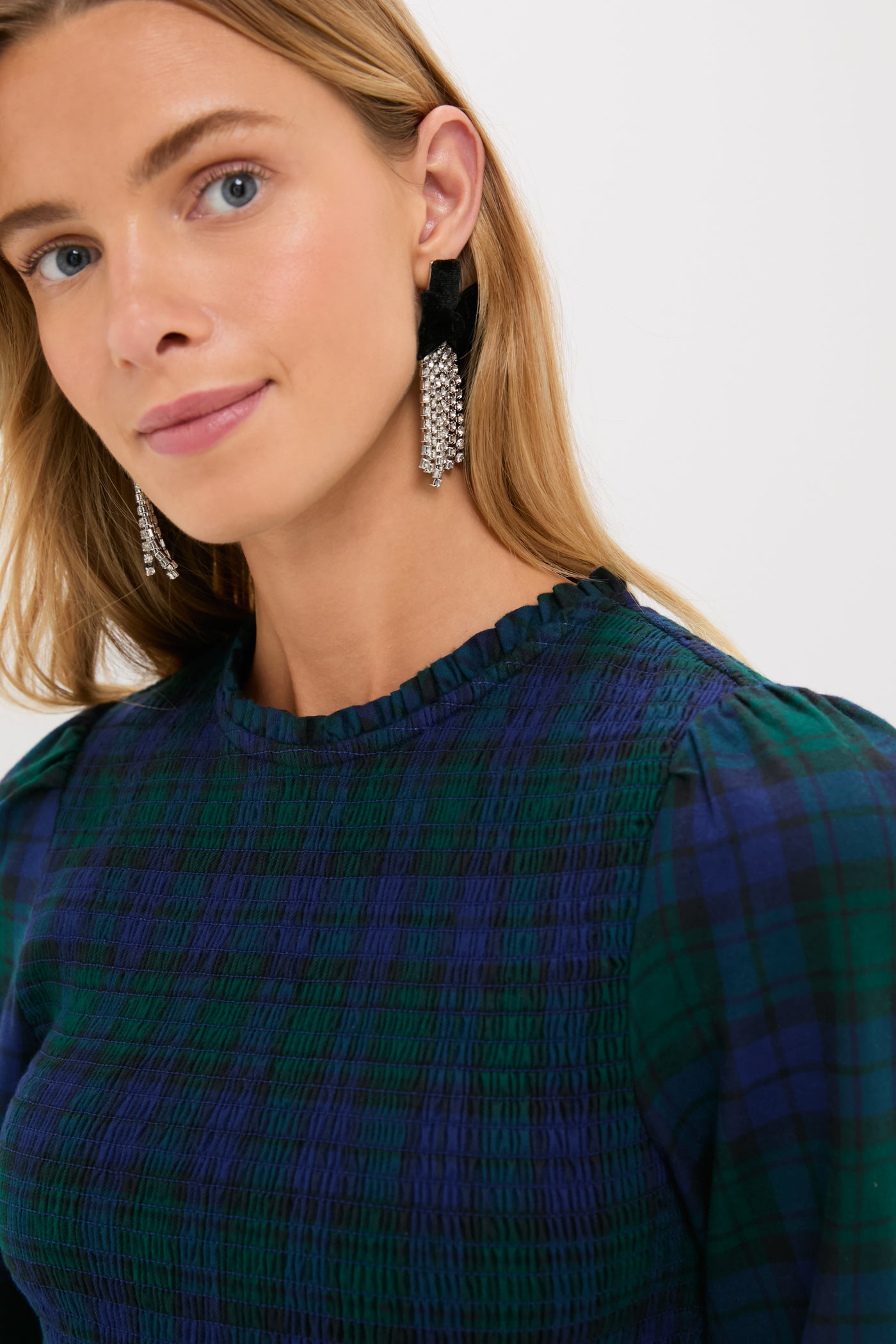 A woman with long blond hair models a green and blue Blackwatch tartan plaid top by Hill House Home, paired with dangling silver earrings, standing against a plain light background.