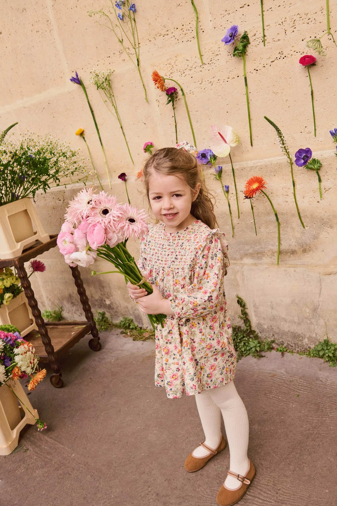 Young girl wearing the classic Cream Wild Flowers Saralou Dress, effortlessly holding a fun bouquet, surrounded by high-end floral decor, embodying playful elegance and charm.