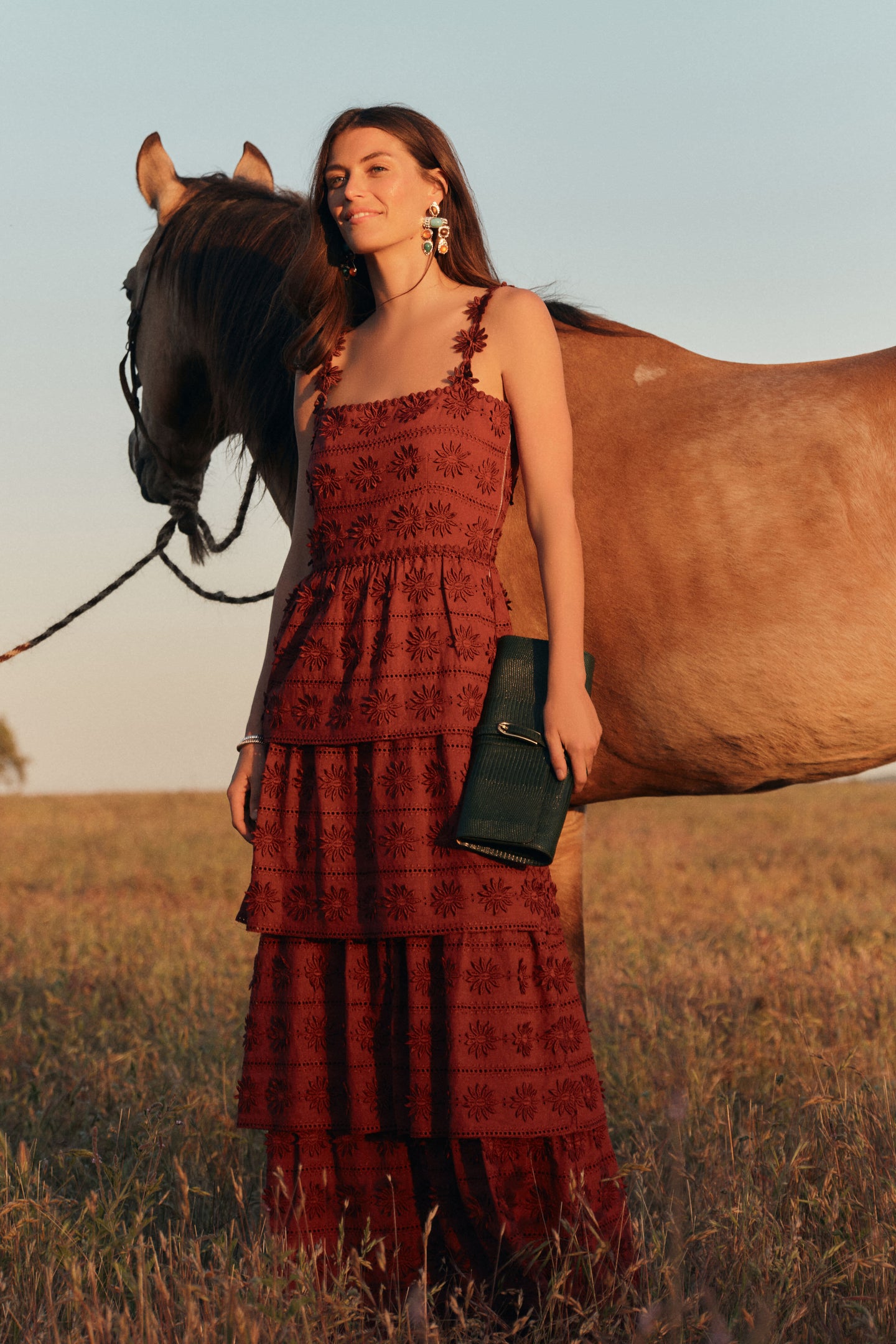 Woman wearing a sleeveless maxi dress with eyelet details in a rust color standing next to a horse. 