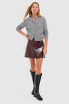 Young woman models the classic Oxblood Meeghan Mini Skirt in glossy vegan leather, paired with a gray cardigan and knee-high boots, showcasing effortless, high-end style.