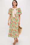 A woman wears the Multi Green Begonia Floral Mimi Midi Dress with a ruffle collar and puff sleeves, layered with a light pink sweater over her shoulders. She holds a woven straw bag and pairs the look with tan sandals.