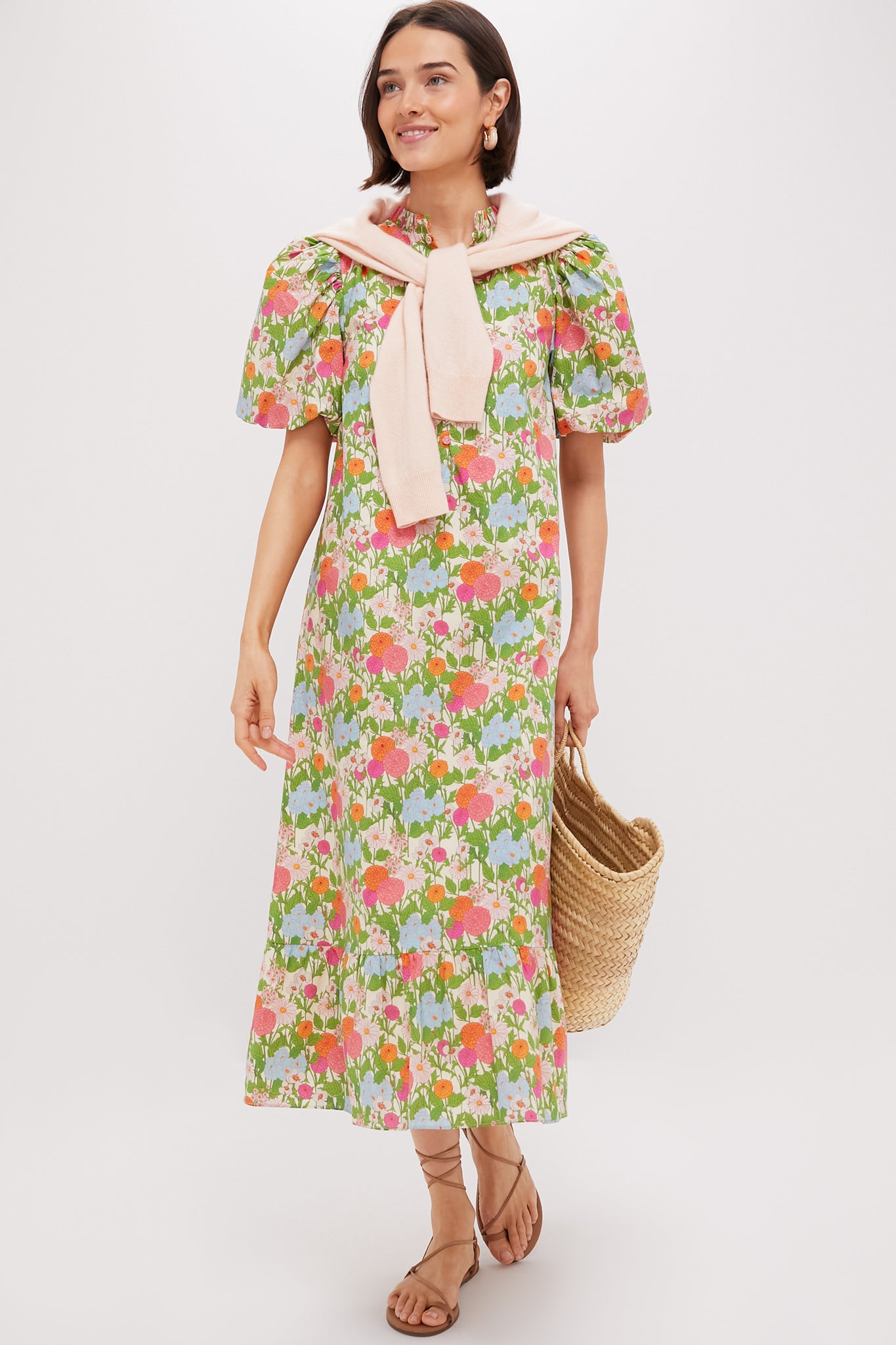 A woman wears the Multi Green Begonia Floral Mimi Midi Dress with a ruffle collar and puff sleeves, layered with a light pink sweater over her shoulders. She holds a woven straw bag and pairs the look with tan sandals.