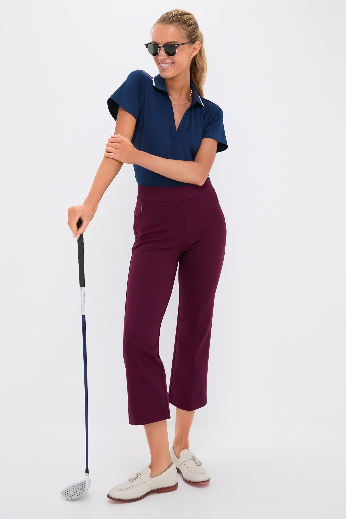 Woman in sporty-casual outfit, showcasing the Petite Winetasting Compression September Pant with a classic, effortless style, holding a golf club against a white backdrop.