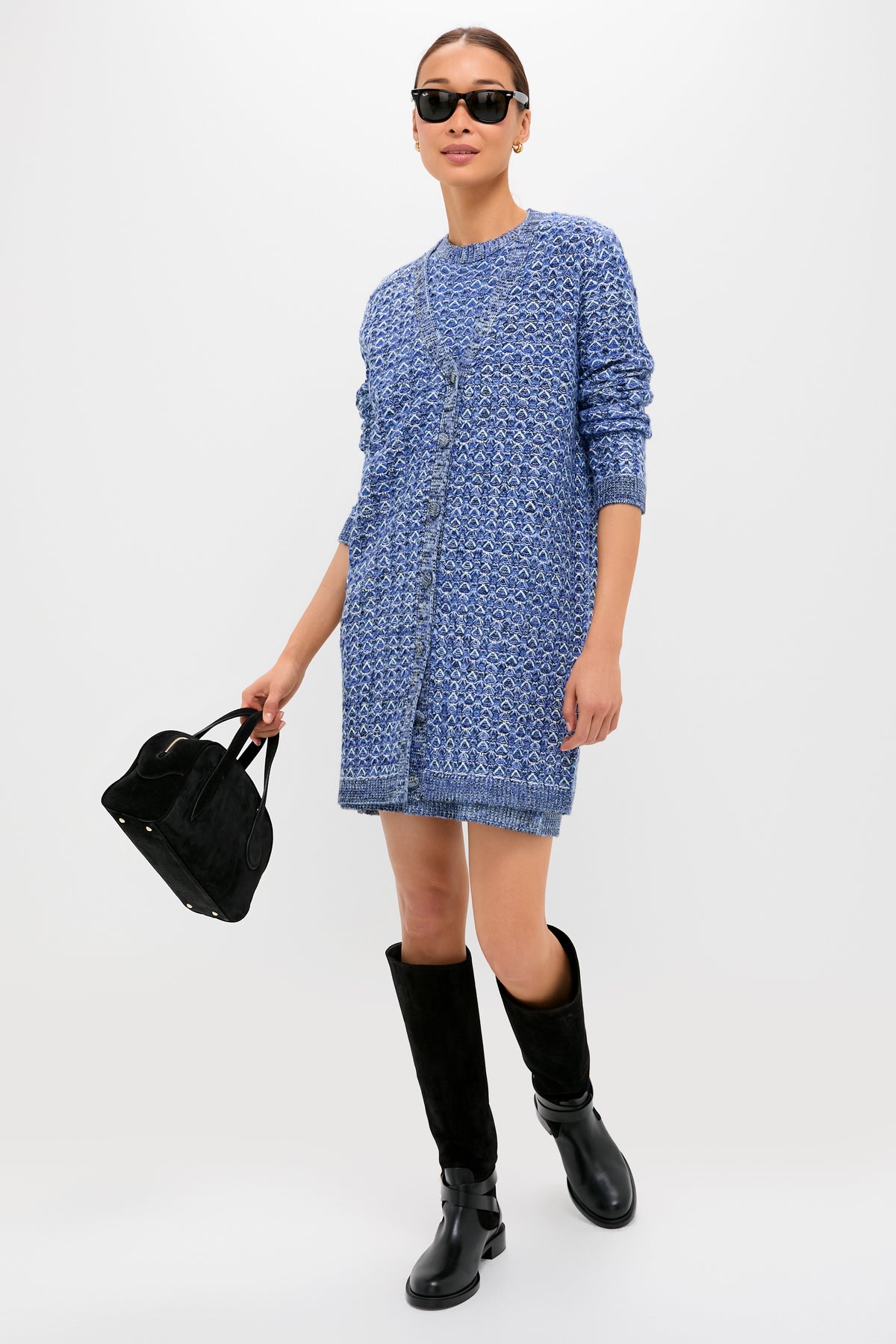 A woman showcases effortless chic in Tuckernuck sunglasses, a blue patterned knit dress, black knee-high boots, and a black handbag, posed against a clean white background.