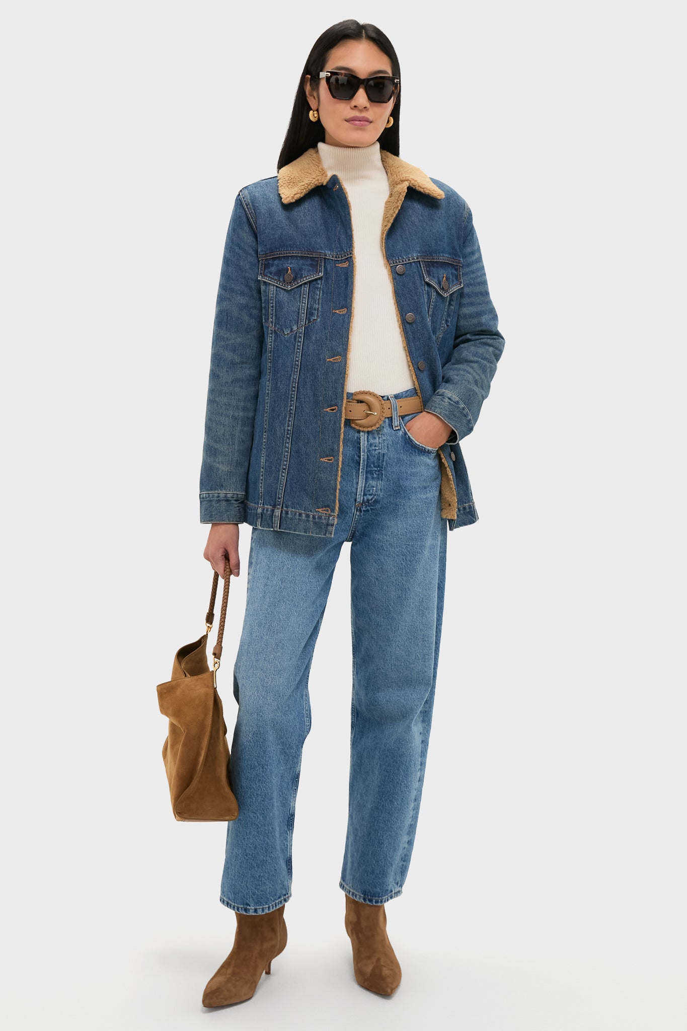 Wearing a Veronica Beard oversized fit denim jacket with sherpa-lined collar, cream turtleneck, blue jeans, brown belt, brown suede boots, sunglasses, and holding a brown suede bag.