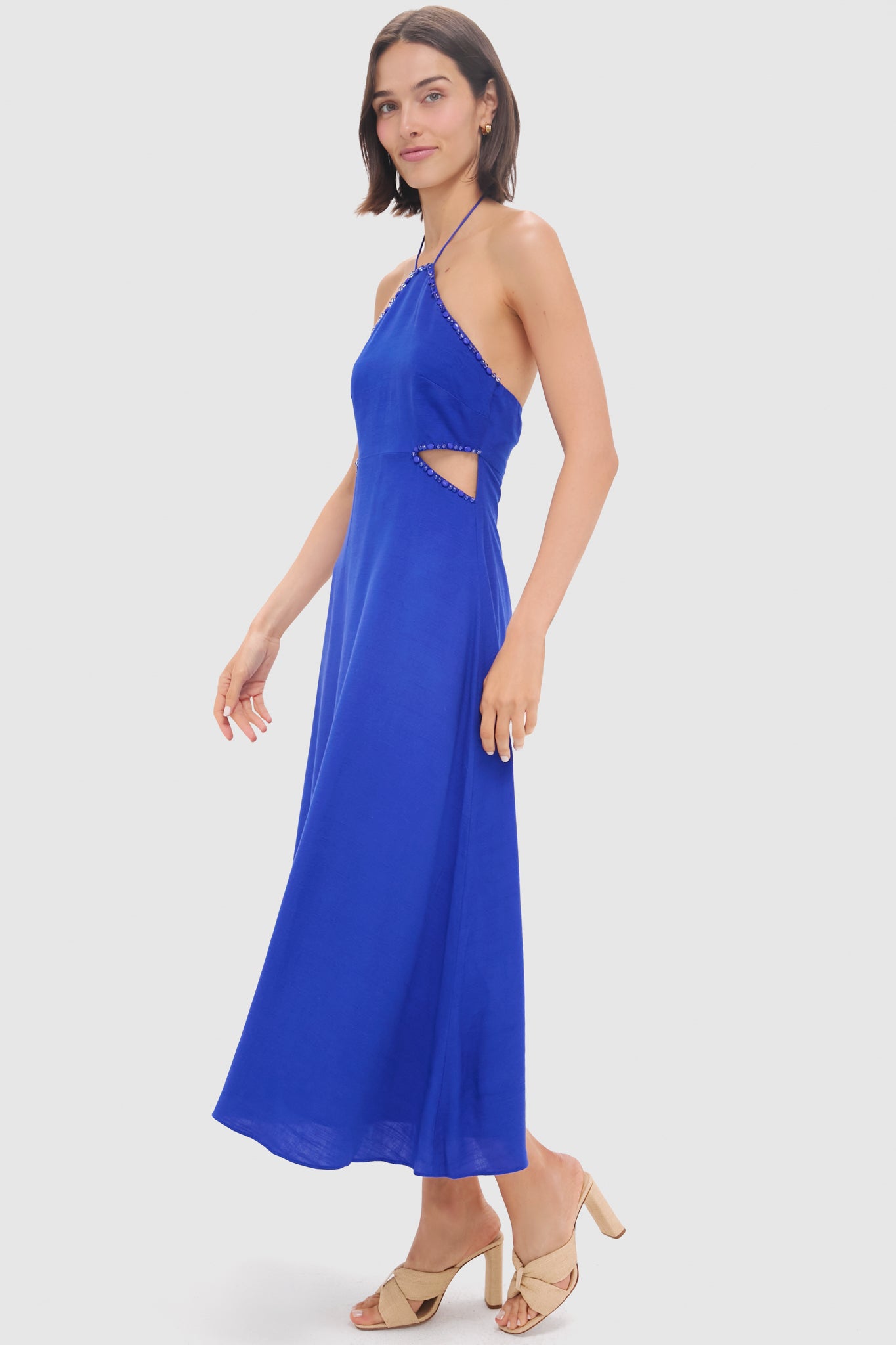 A woman stands against a plain light background in a Hyacinth House Royal Blue Beaded Azure Midi Dress with side cutout detail and beige heeled sandals—a flirty, bohemian piece ideal for summer occasions.