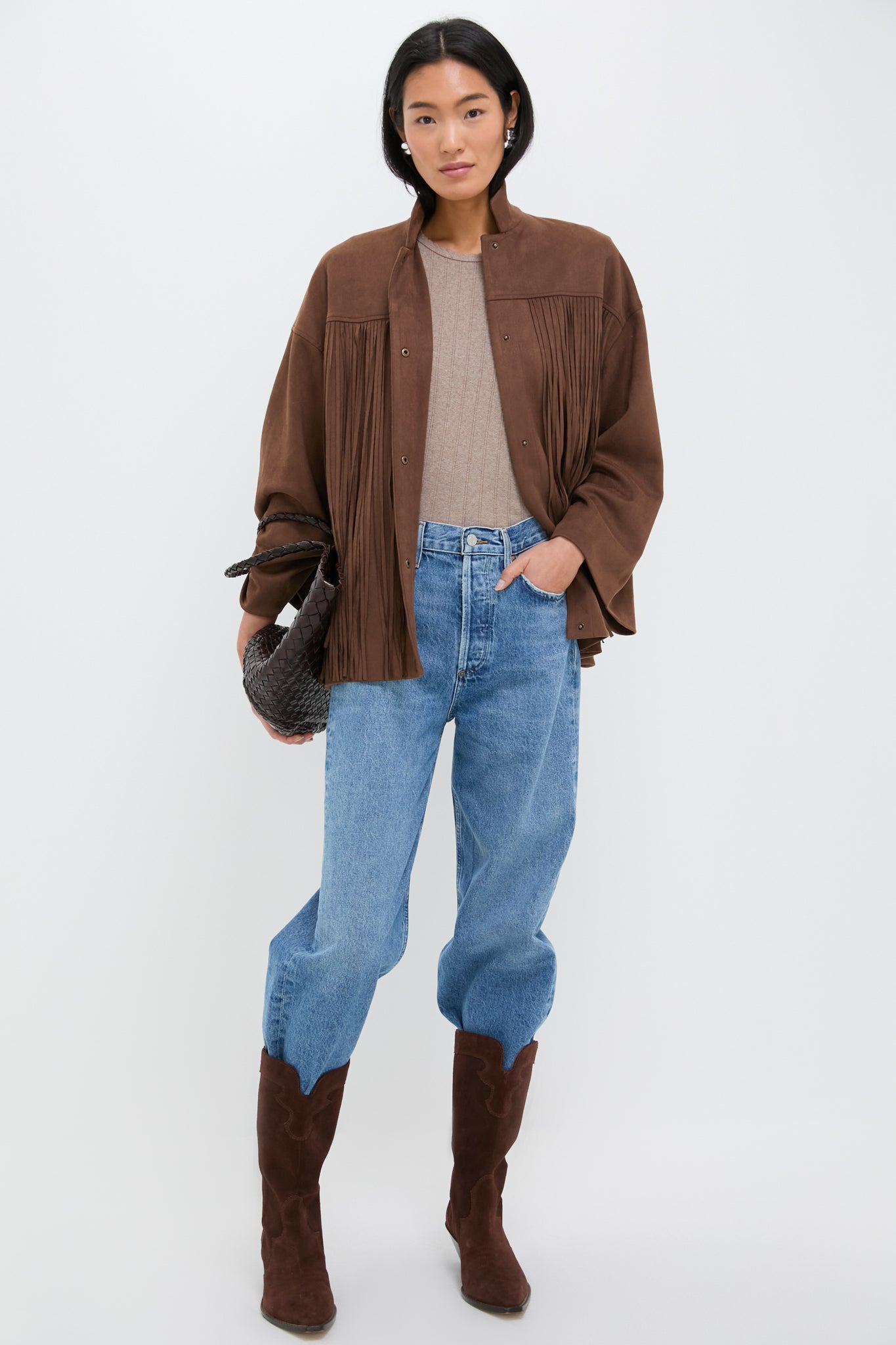 A woman wears a Tuckernuck chocolate suede jacket over a beige top, blue jeans, and brown suede boots, carrying a woven bag.