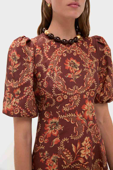 Woman shown from the shoulders up wears an Alemais brown transition midi dress with orange floral patterns and puff sleeves, accessorized with a chunky beaded necklace.