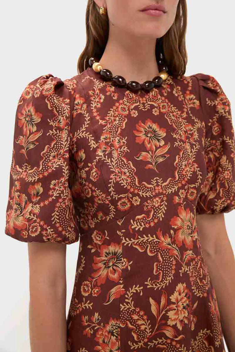Woman shown from the shoulders up wears an Alemais brown transition midi dress with orange floral patterns and puff sleeves, accessorized with a chunky beaded necklace.