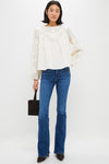 A woman stands against a white background wearing a Tuckernuck relaxed fit 100% cotton embroidered blouse, blue bootcut jeans, brown pointed shoes, and holding a small black handbag.