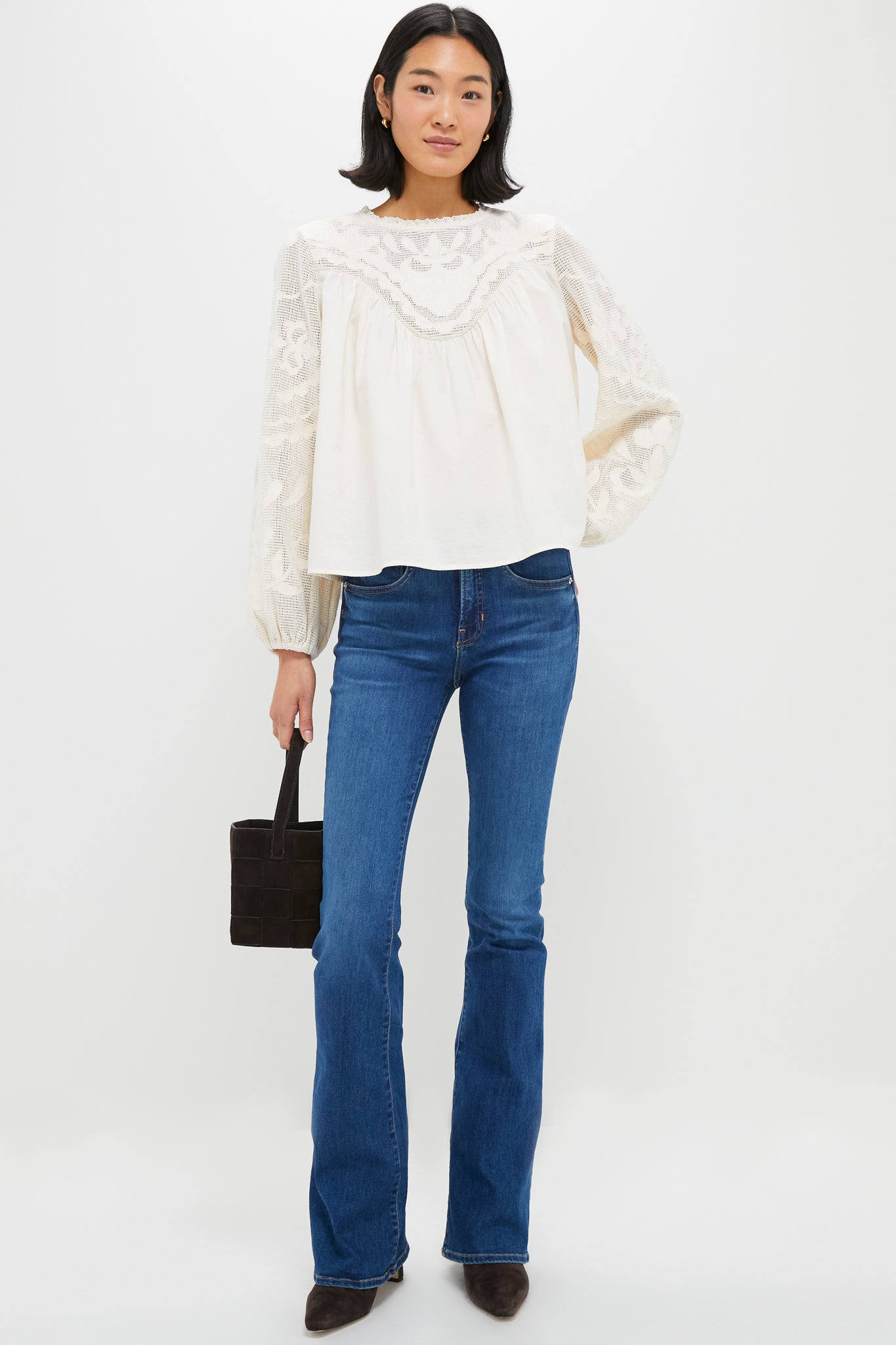 A woman stands against a white background wearing a Tuckernuck relaxed fit 100% cotton embroidered blouse, blue bootcut jeans, brown pointed shoes, and holding a small black handbag.