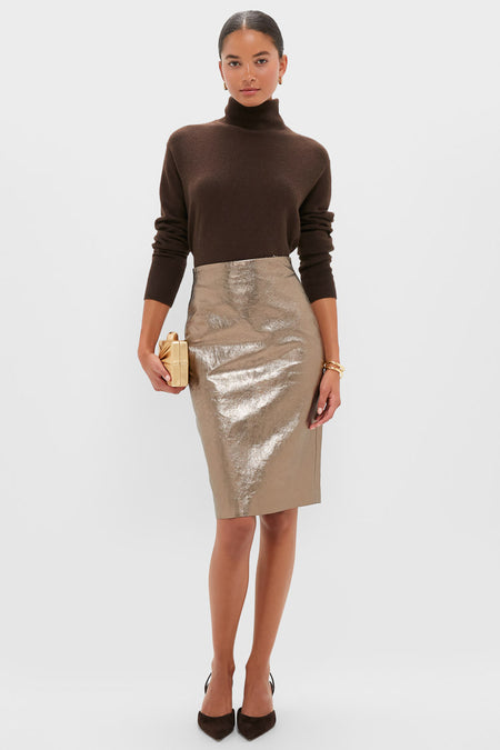 A woman models refined women’s clothing by Andres Otalora: an elegant brown turtleneck sweater, metallic gold pencil skirt, dark pointed heels, and a gold clutch, all set against a plain white background.