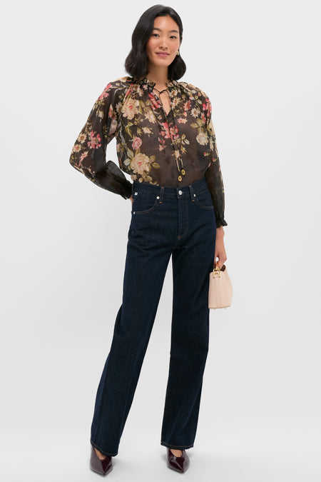 A woman stands against a white background wearing a sheer floral blouse, dark blue jeans, and Marc Fisher burgundy pointed-toe kitten heels, holding a small beige handbag.
